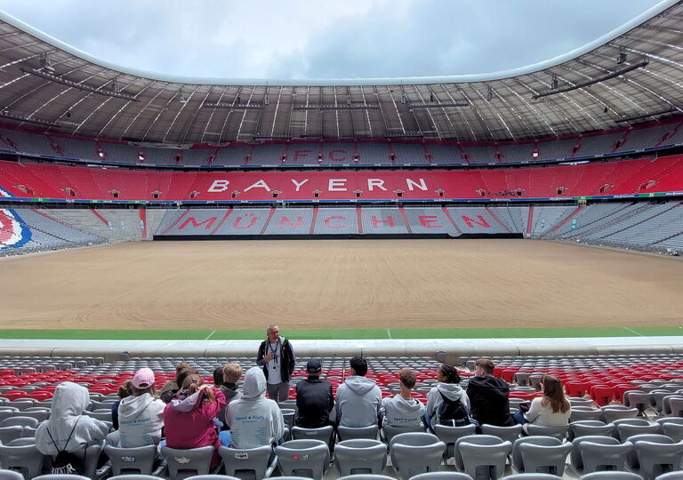 Schüler sitzen im Stadion des FC Bayern München und bekommen Erklärungen von einem Guide