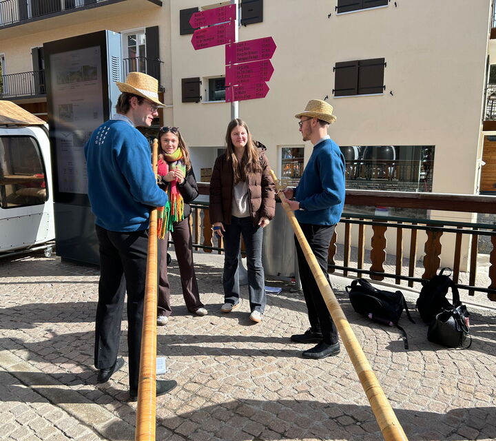 Zwei Schülerinnen unterhalten sich mit zwei Männern in blauen Pullovern, die Alphörner in der Hand haben