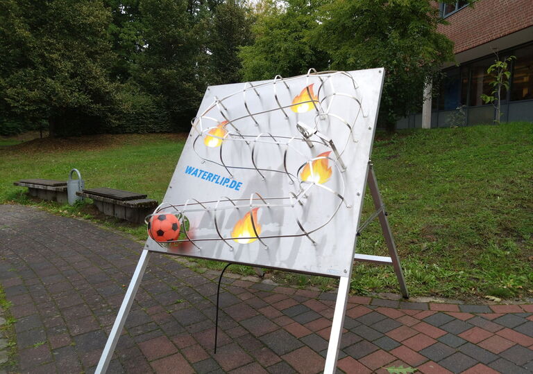 Bild einer Wand, an der ein roter Ball mithilfe eines Wasserschlauchs nach oben gespritzt werden muss