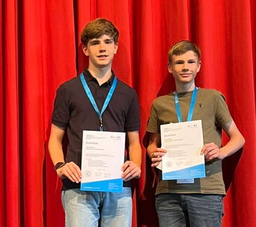 Jannik und Felix stehen vor einem roten Vorhang und halten ihre Urkunde in der Hand