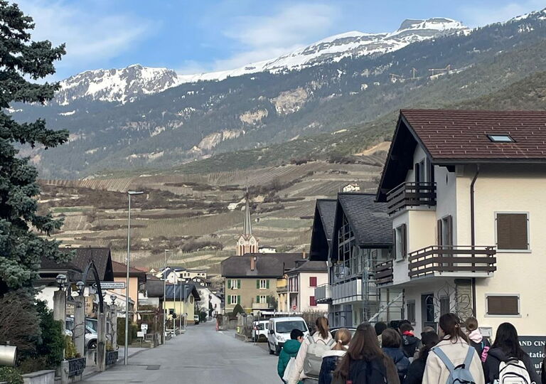 Eine Schülergruppe läuft auf der rechten Seite an einer Alpenstraße entlang. Im Hintergrund sieht man die Schweizer Alpen.