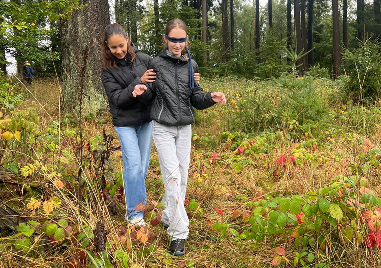 Eine Schülerin führt eine andere Schülerin mit verbundenen Augen durch den Wald