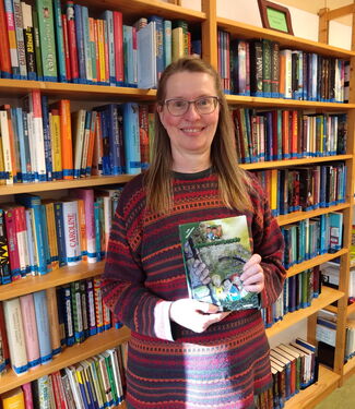 Frau Brauer mit einem Buch in der Hand vor einem Bücherregal stehend