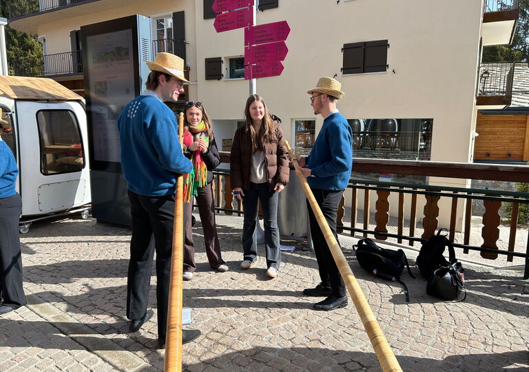 Zwei Schülerinnen unterhalten sich mit zwei Männern in blauen Pullovern, die Alphörner in der Hand haben
