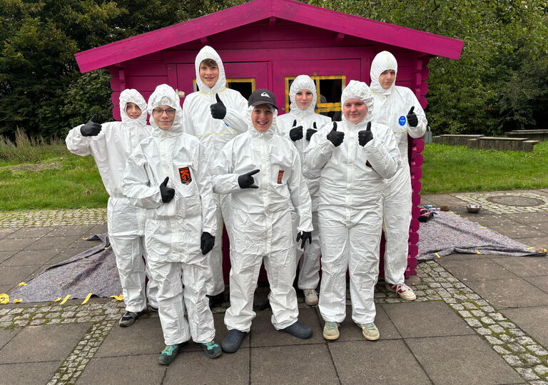 Sieben Schülerinnen und Schüler in weißen Maleranzügen im Gruppenfoto vor der pinken Spielehütte